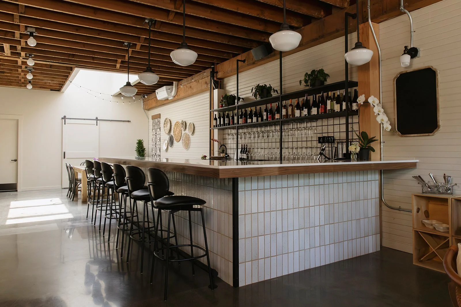 Photo of a modern bar interior with high chairs along a curved counter, hanging pendant lights, a wall with decorative plates, and shelves with wine bottles and glasses.