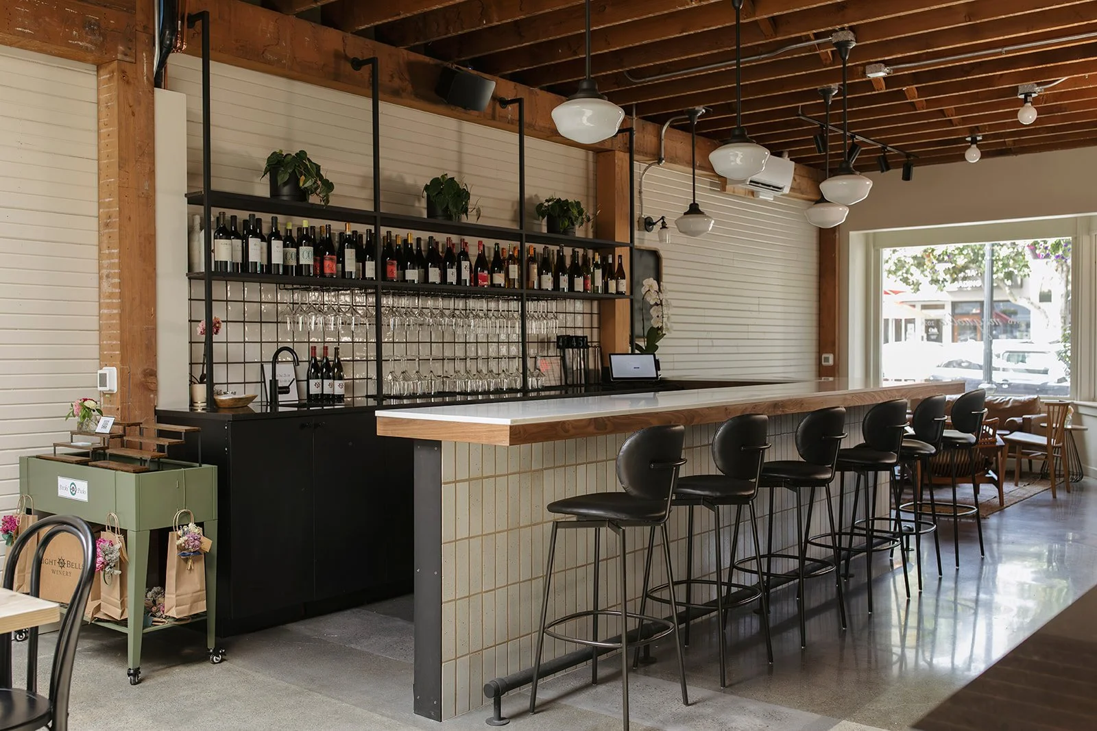Interior of a modern cafe or bar with a row of black bar stools along a white-tiled counter, wine bottles and glasses displayed on shelves behind the counter, large window with natural light, and wooden ceiling with hanging lights.