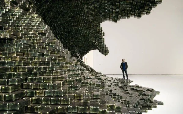 A person standing in an art installation resembling a large wave made of metallic blocks in a white gallery space.