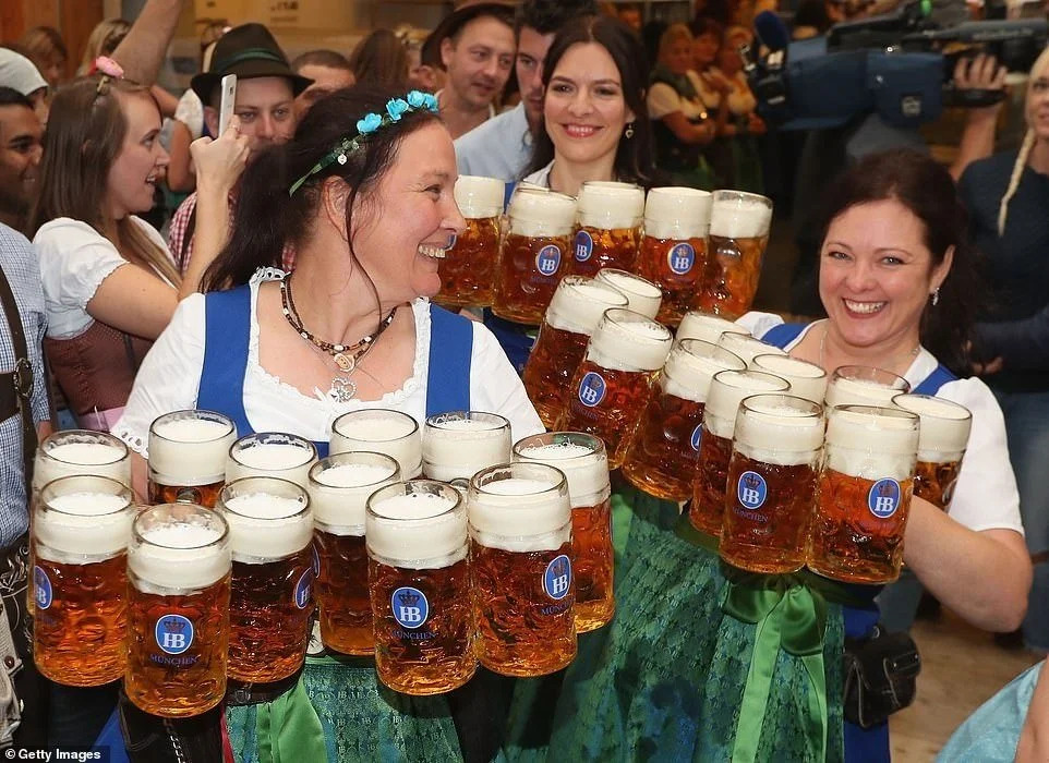 Women wearing traditional Dirndl dresses celebrating with large beer steins at a lively event, likely Oktoberfest.