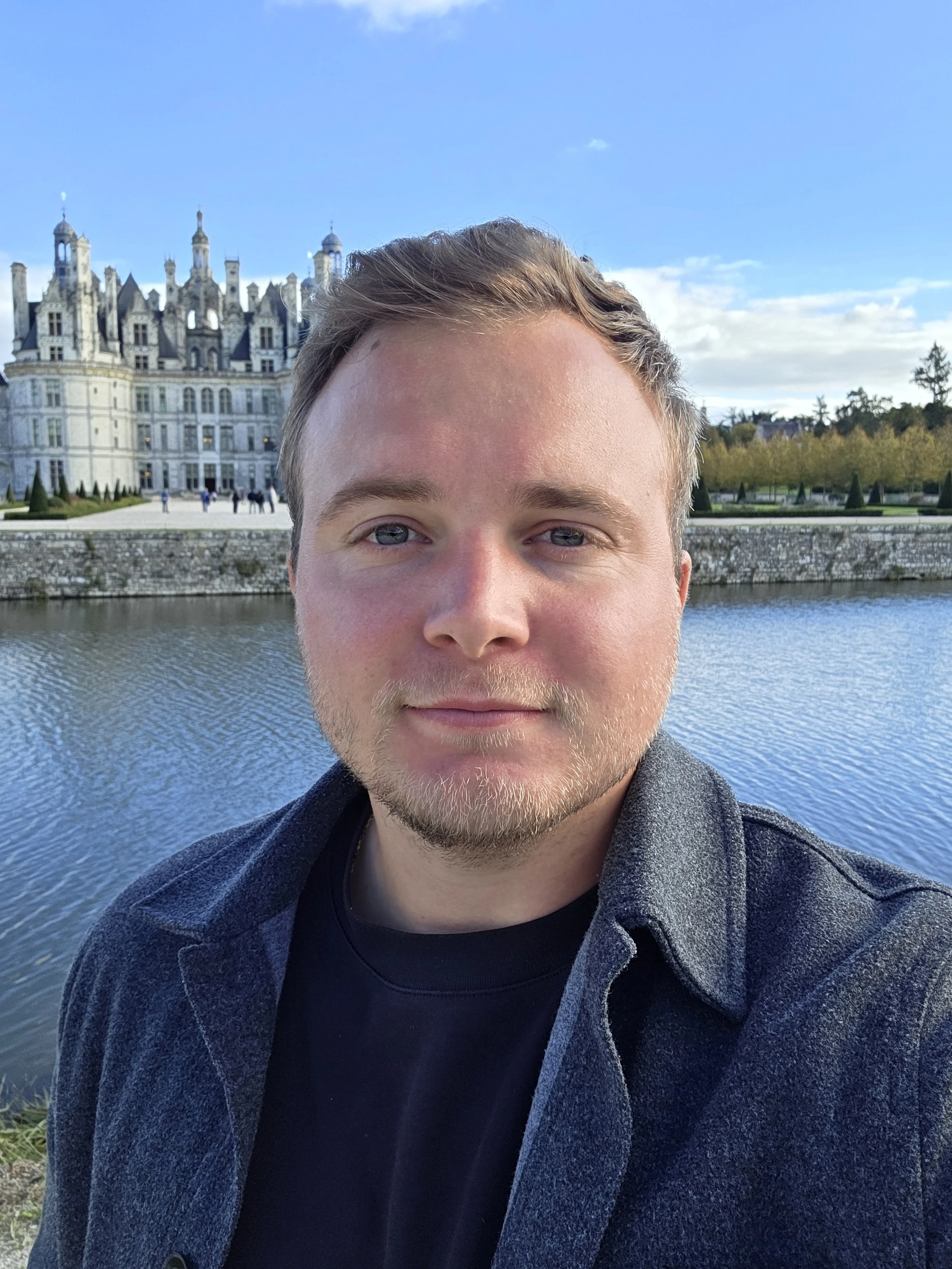 A young man taking a selfie near a body of water with a large, castle-like building in the background during daytime.