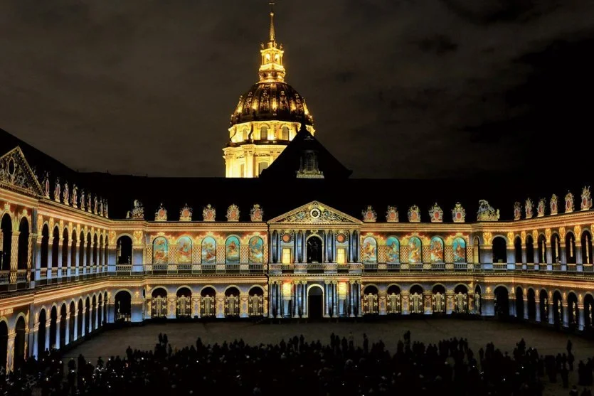 Night view of a historic building with illuminated frescoes and a central dome illuminated by golden light, with cloudy sky in the background.