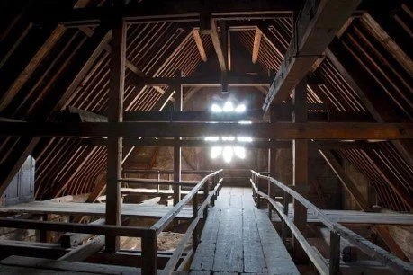 View of the attic interior with wooden beams and a walkway, illuminated by light coming through small windows.