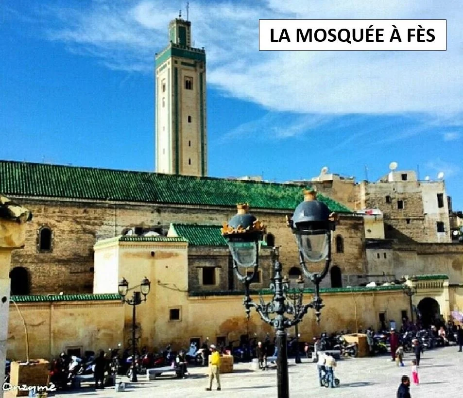 Scene of a historic mosque with beige stone walls and green roofs, under a bright blue sky, with people, motorcycles, and cars in a busy square.