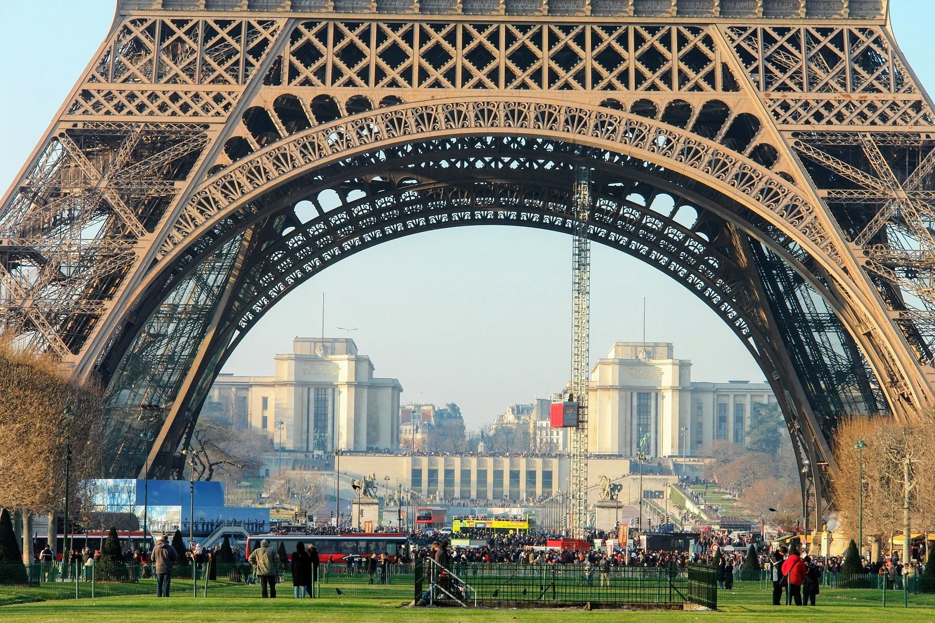 Looking up at the Eiffel Tower in Paris, France, with a landscaped park and people in the foreground, and buildings and a clear sky in the background.
