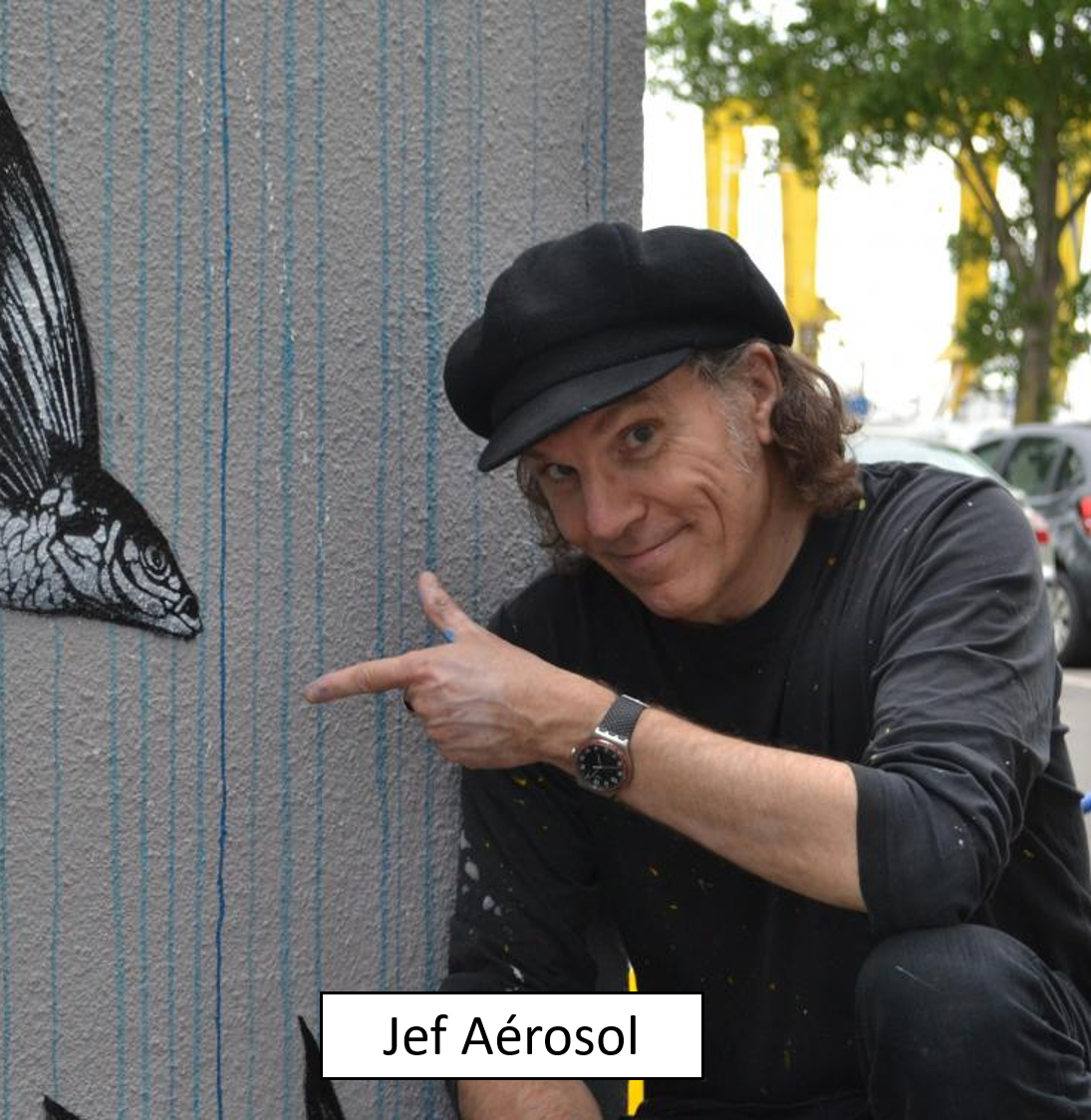 A man in black clothing and a black cap pointing at a wall with a black and white butterfly mural, standing outdoors with cars and trees in the background.