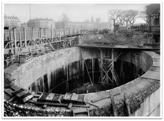 Black and white photo of a large construction site with a deep, round excavation and wooden scaffolding. There are trees and buildings in the background.