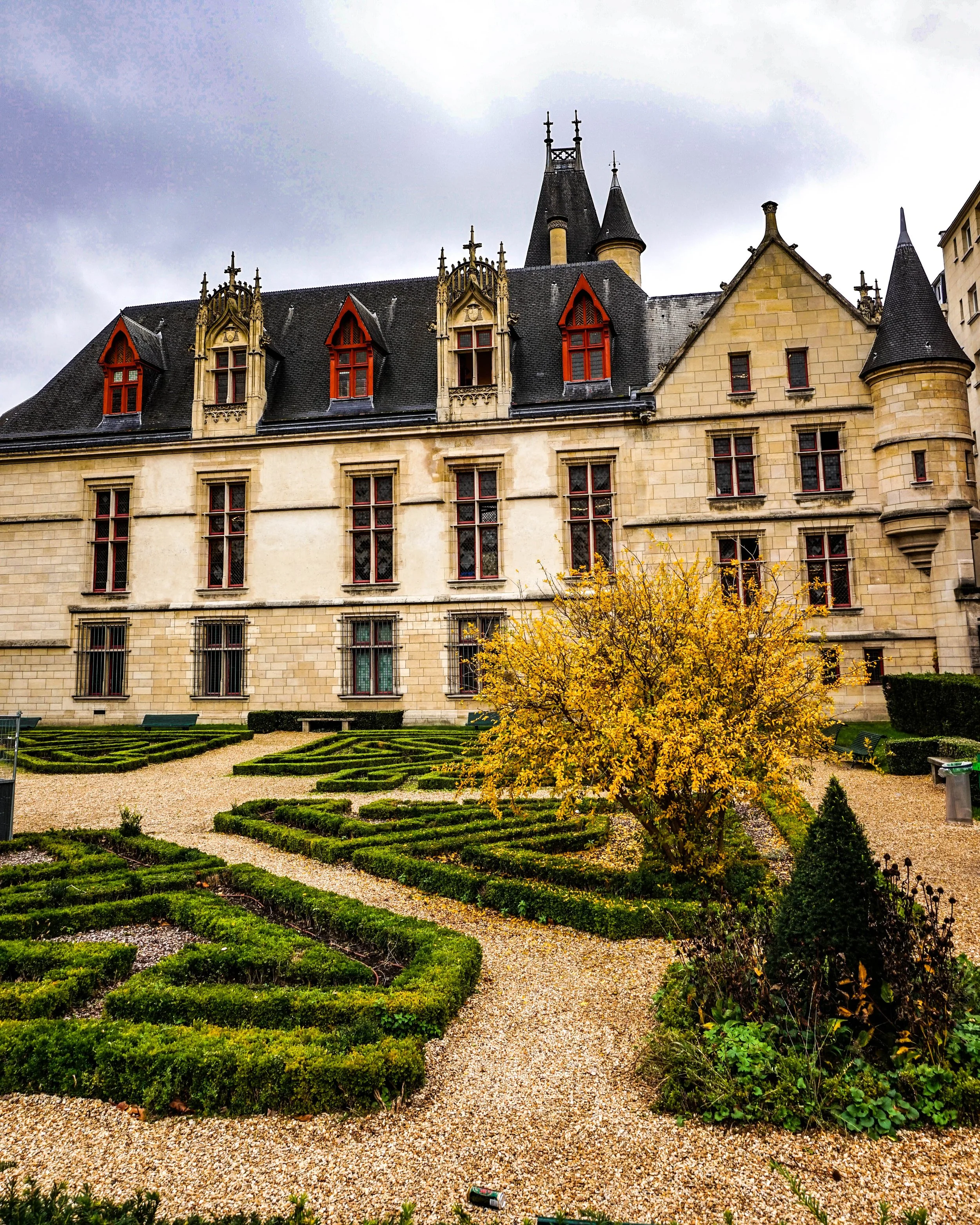 A historic stone castle with pointed roofs and red window frames, surrounded by a neatly manicured garden with a yellow-leaved tree and gravel pathways, under a cloudy sky.