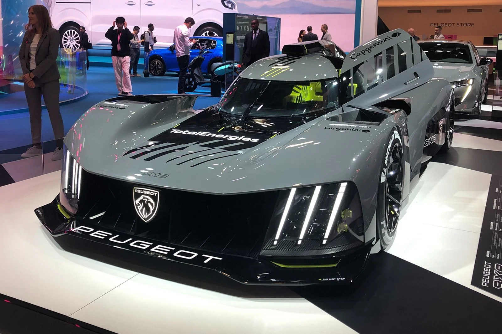 A sleek, futuristic Peugeot race car on display at an auto show, featuring a gray and black exterior with sharp angles, LED headlights, and car branding including 'Peugeot' and 'TotalEnergiers.' Several people are in the background, observing other vehicles and exhibits.