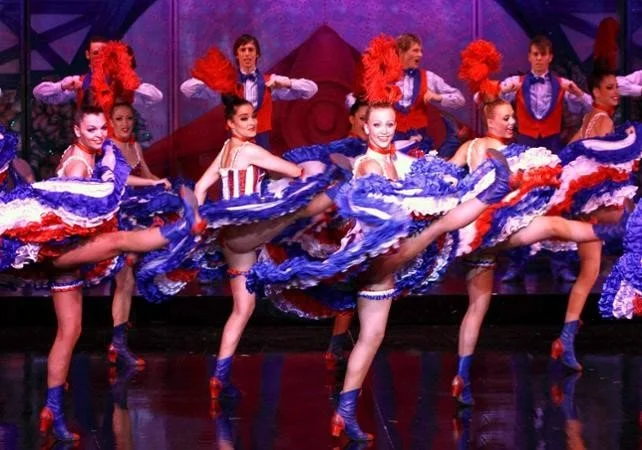 Group of dancers in colorful costumes performing on stage, with girls lifting their skirts and boys standing behind them.