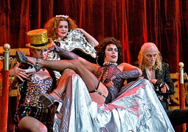 Four performers in colorful costumes and makeup posing on a stage with a red curtain, some sitting on a throne-like chair and others reclining or sitting on the floor.