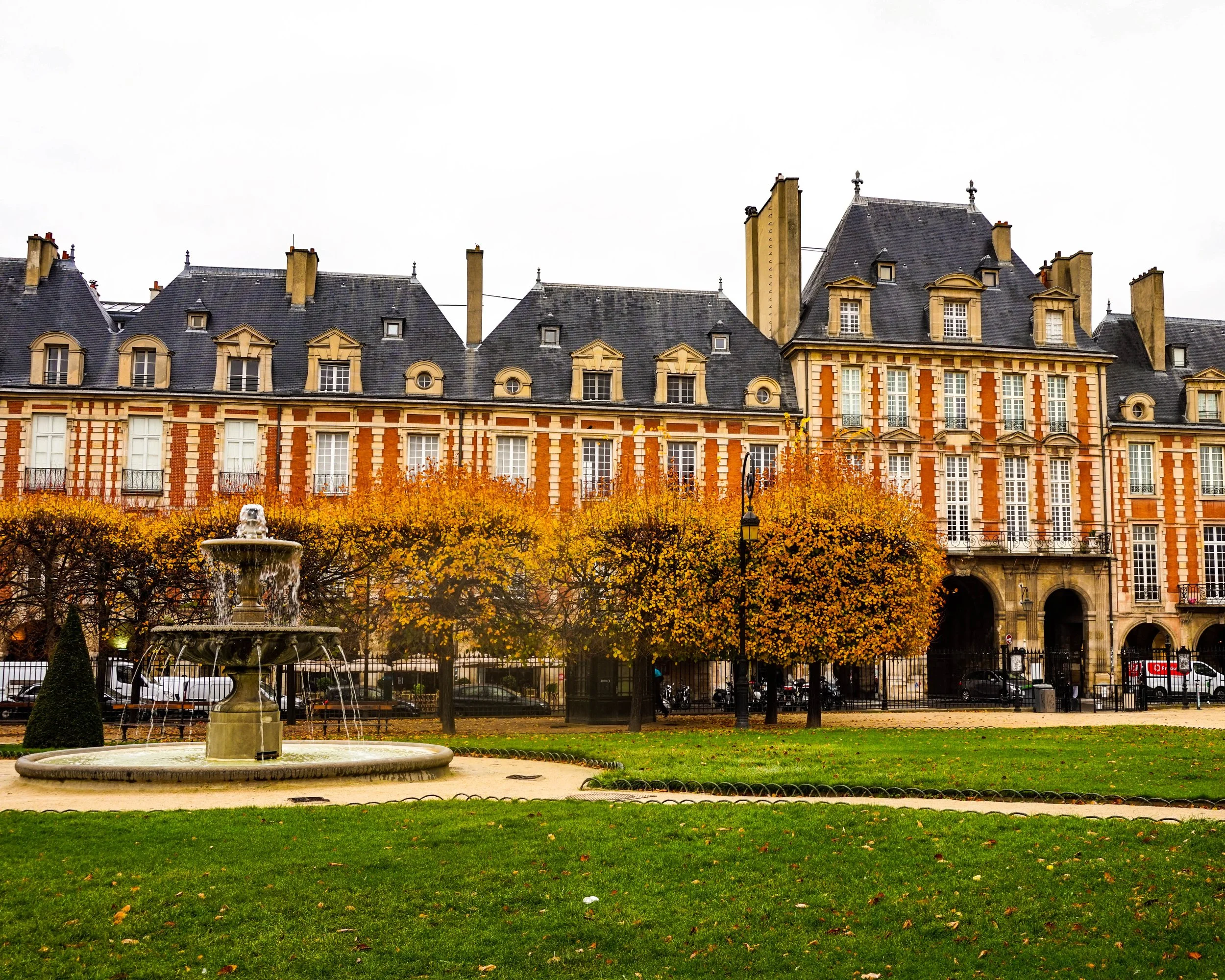 European-style building with red brick and white accents, surrounded by trees with yellow leaves, in a park with a fountain in the foreground.