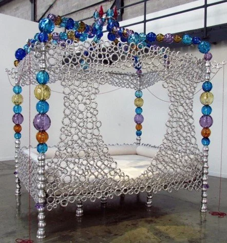 A bed designed to look like a giant dreamcatcher with rings and bead decorations, set in an art gallery.