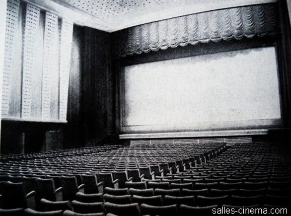 Empty movie theater with rows of seats facing a large screen and curtains in the background.