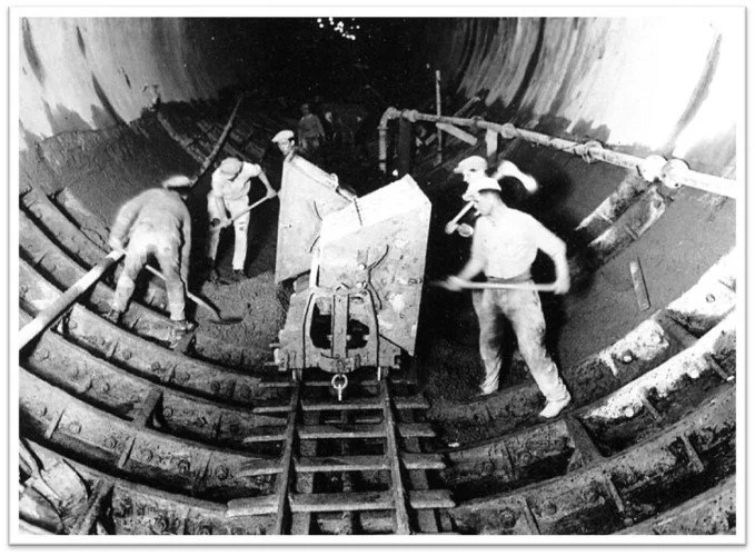 Workers in a large tunnel construction site are installing a large piece of equipment using tools and scaffolding.