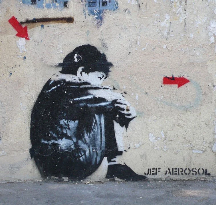 Street art stencil of a person sitting with their knees pulled up, resting their face on their arms, on a weathered wall. Two red arrows point to the left and right from the figure. The names "JEB" and "AEROSOL" are written at the bottom.