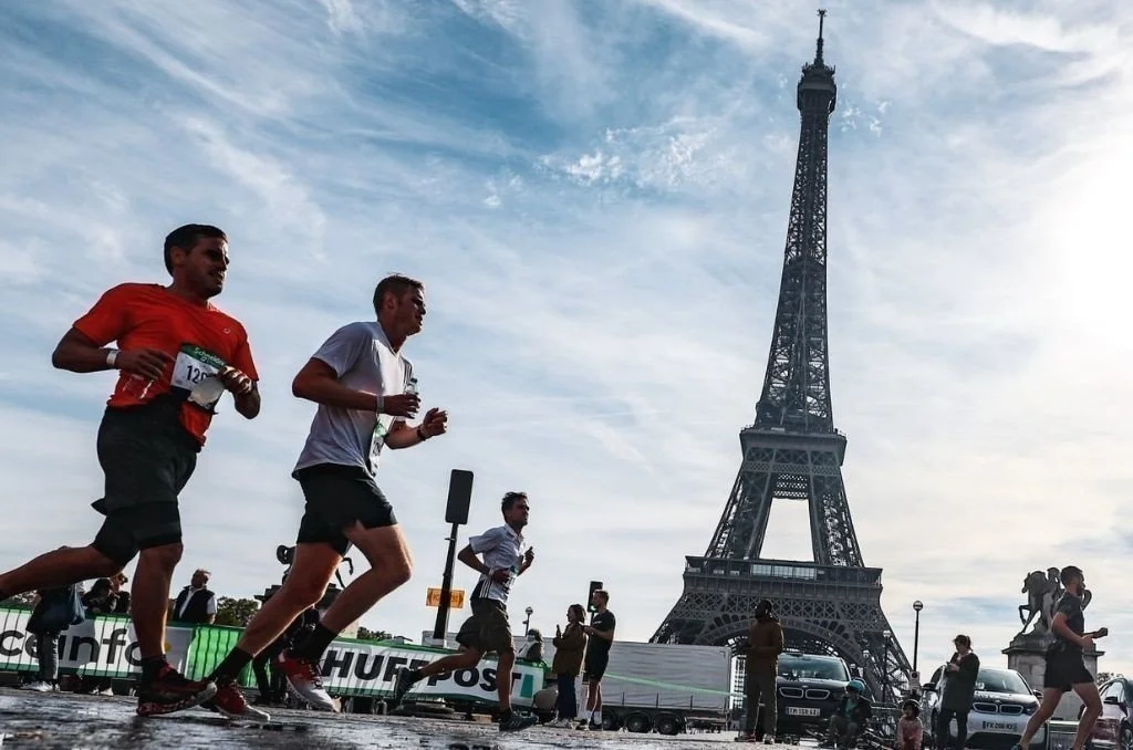Three men running marathons near the Eiffel Tower in Paris, France, with spectators and vehicles in the background.