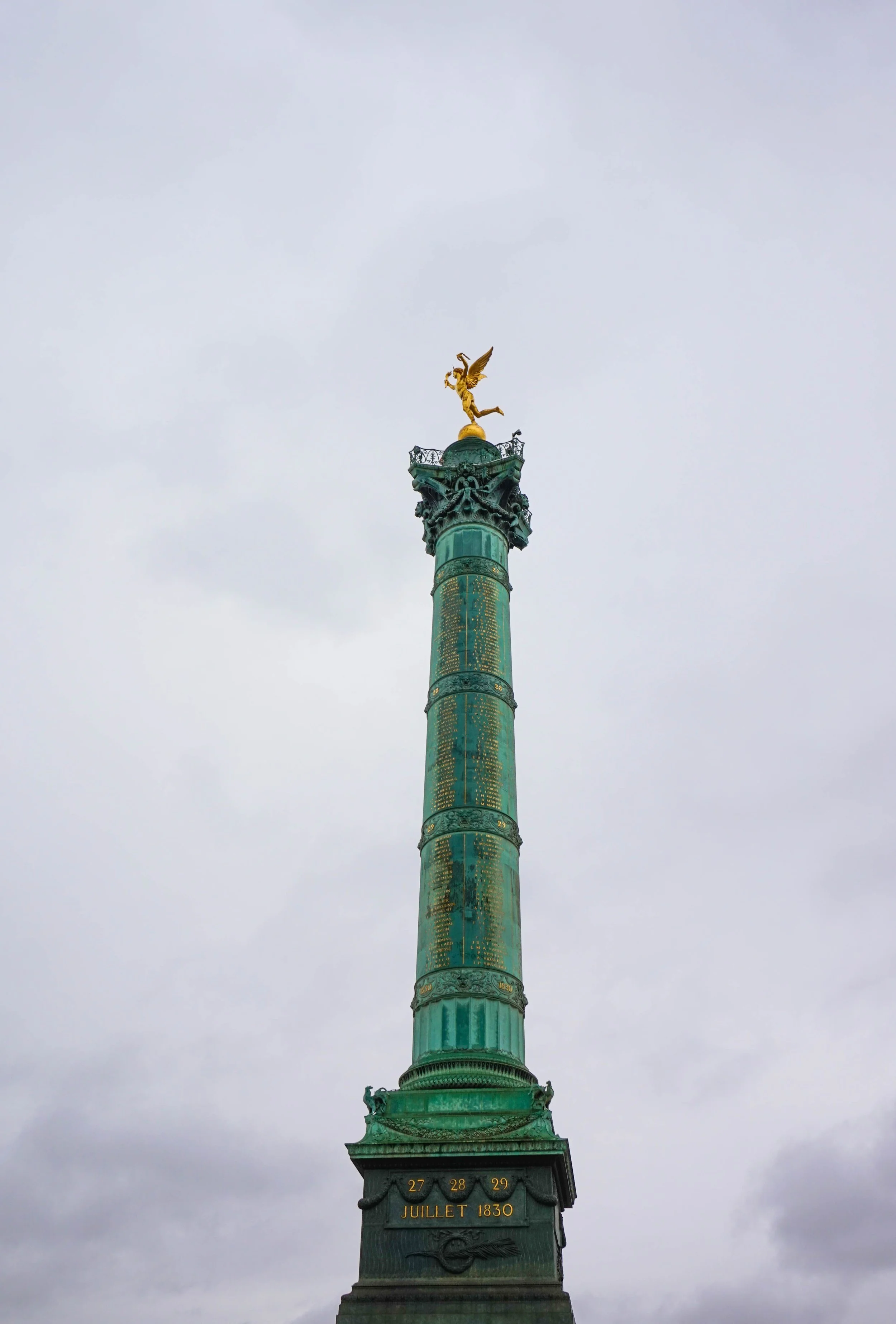 A tall green monument with a golden statue of a winged figure on top, against a cloudy sky.