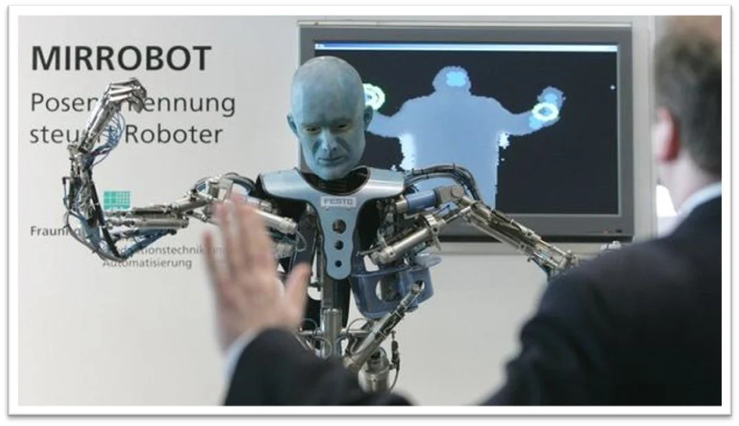 A humanoid robot with a human mask on its face, giving a high-five to a person in a suit. There is a computer screen in the background displaying a digital representation of the robot's body and arms.