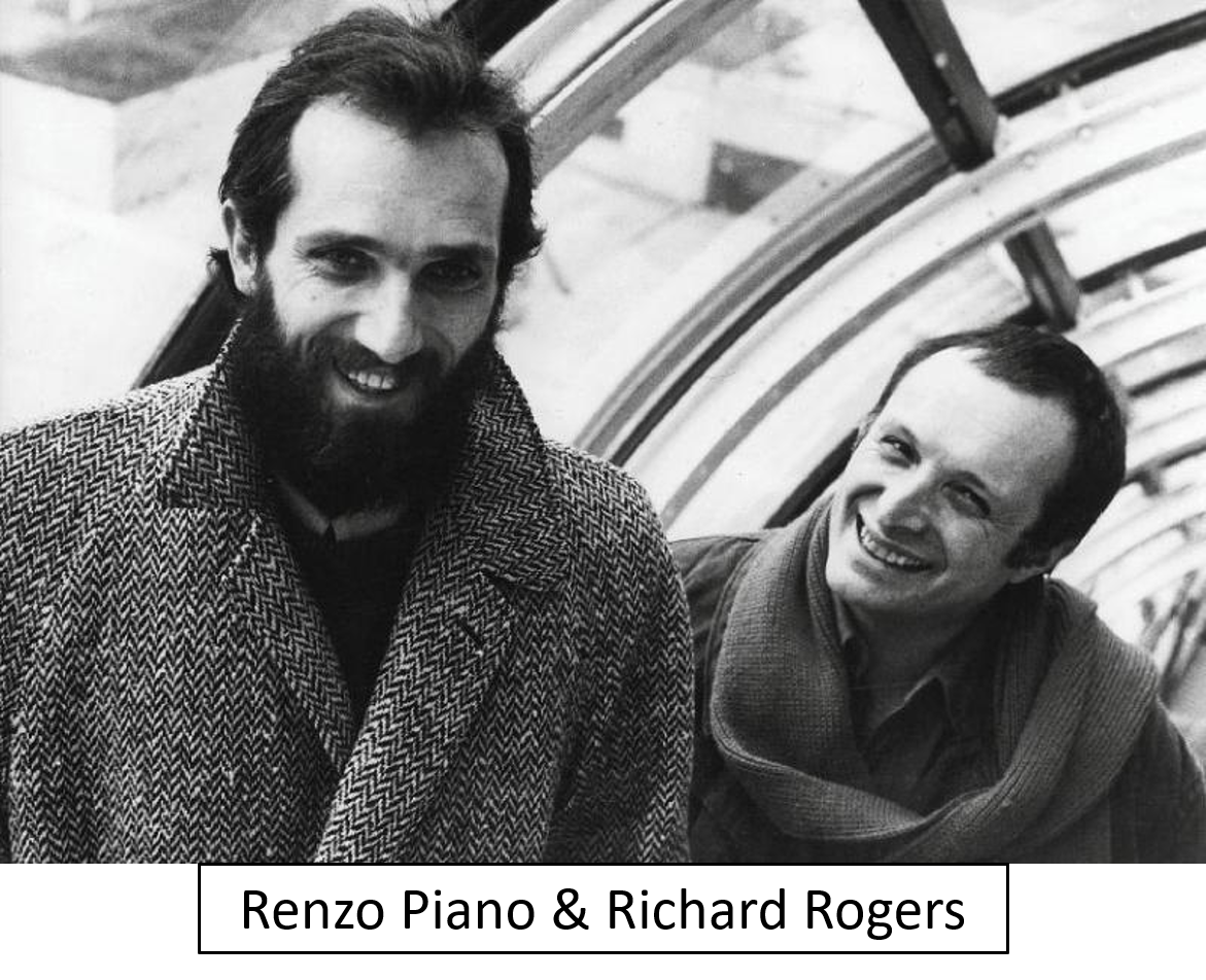 Black and white photograph of two men smiling indoors. The man on the left has a beard and is wearing a patterned coat, while the man on the right is clean-shaven and wearing a jacket and scarf.