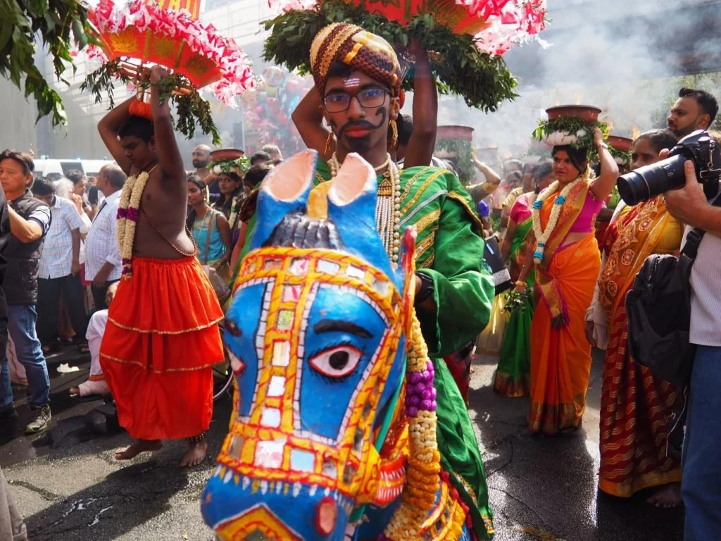 A person dressed in traditional Indian attire riding a decorated horse costume during a cultural parade, with others in colorful clothing participating, and a crowd watching.
