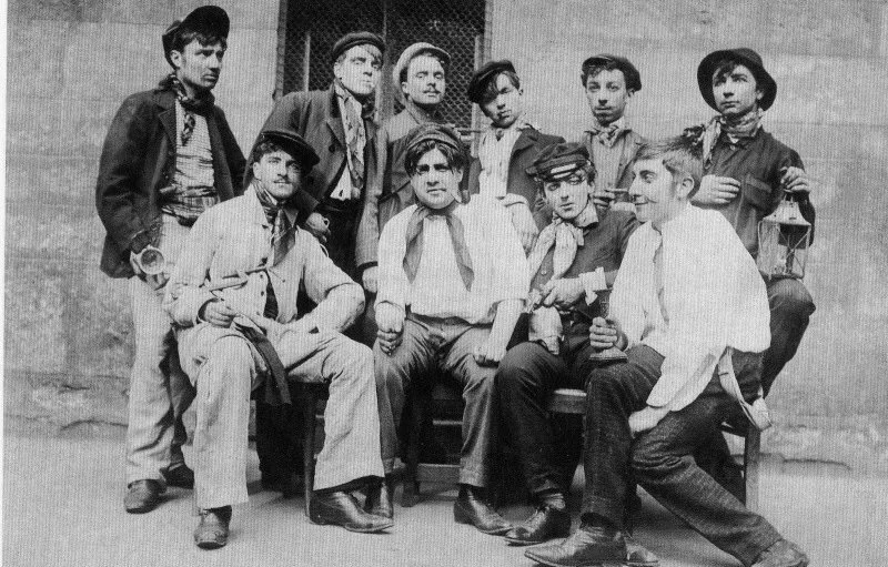 Black and white photo of eleven boys, some seated and some standing, dressed in old-fashioned clothing, posing outdoors against a textured wall.