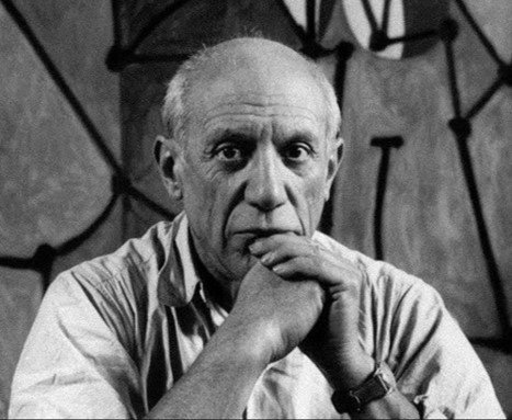 A black and white photograph of an elderly man with short hair, resting his chin on his hand and looking directly at the camera, with climbing ropes in the background.