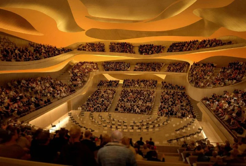 Interior view of a large, modern auditorium filled with an audience seated in tiered rows, with a stage at the front decorated with chairs and music stands, under warm yellow lighting.