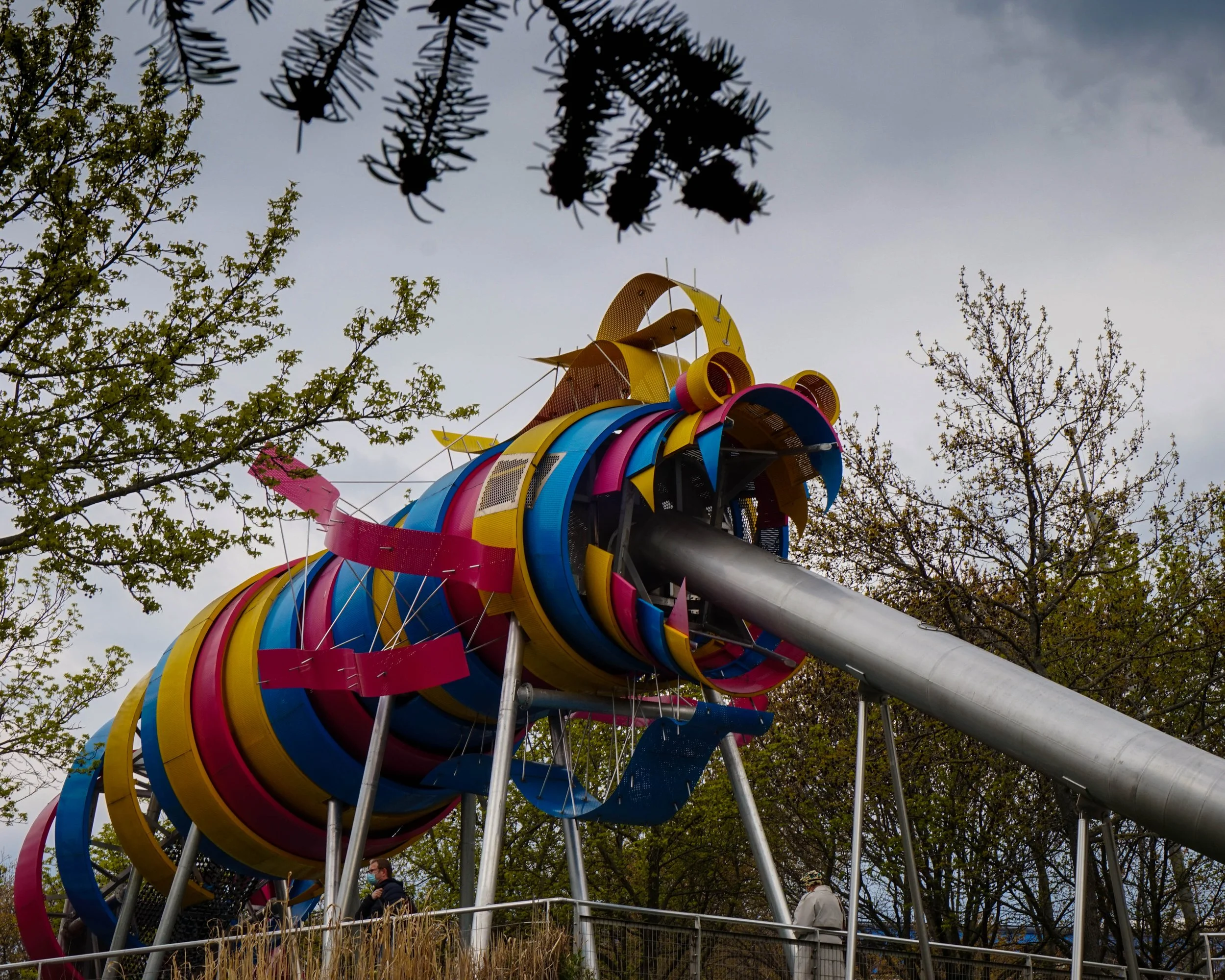 Colorful playground slide with multiple sections made of metal and plastic, set outdoors among trees on a cloudy day.