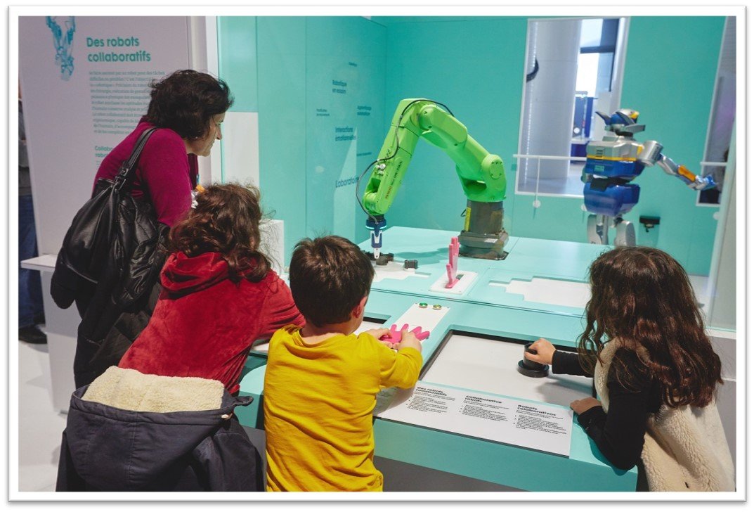 A group of children and an adult observe and interact with robotic arms behind a glass display at a science or technology exhibit.