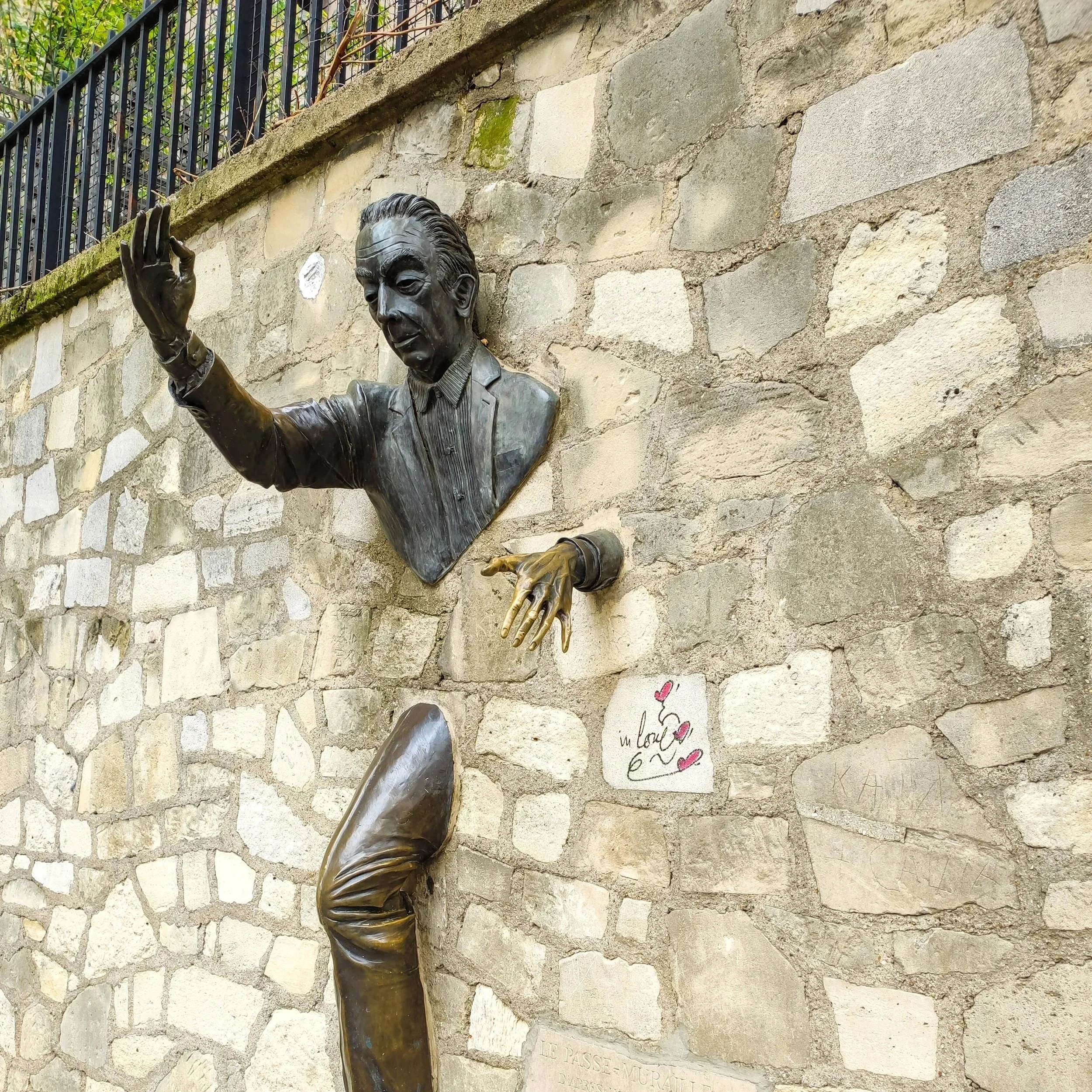 Bronze sculpture of a man emerging from a stone wall, with one hand raised and the other hand pointing downward, along with a partial bronze figure of a leg, on a brick wall with graffiti and a plaque.