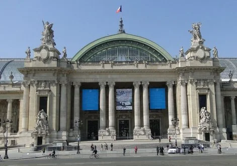 Facade of a historic building with grand columns, statues, and a large arched window, featuring a flag on top.