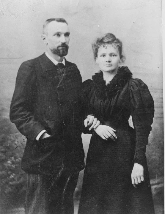 A black-and-white vintage photograph of a man and woman posing together, with the man having a beard and wearing a suit, and the woman with styled hair and wearing a dark dress with puffed sleeves, in front of a blurred outdoor background.
