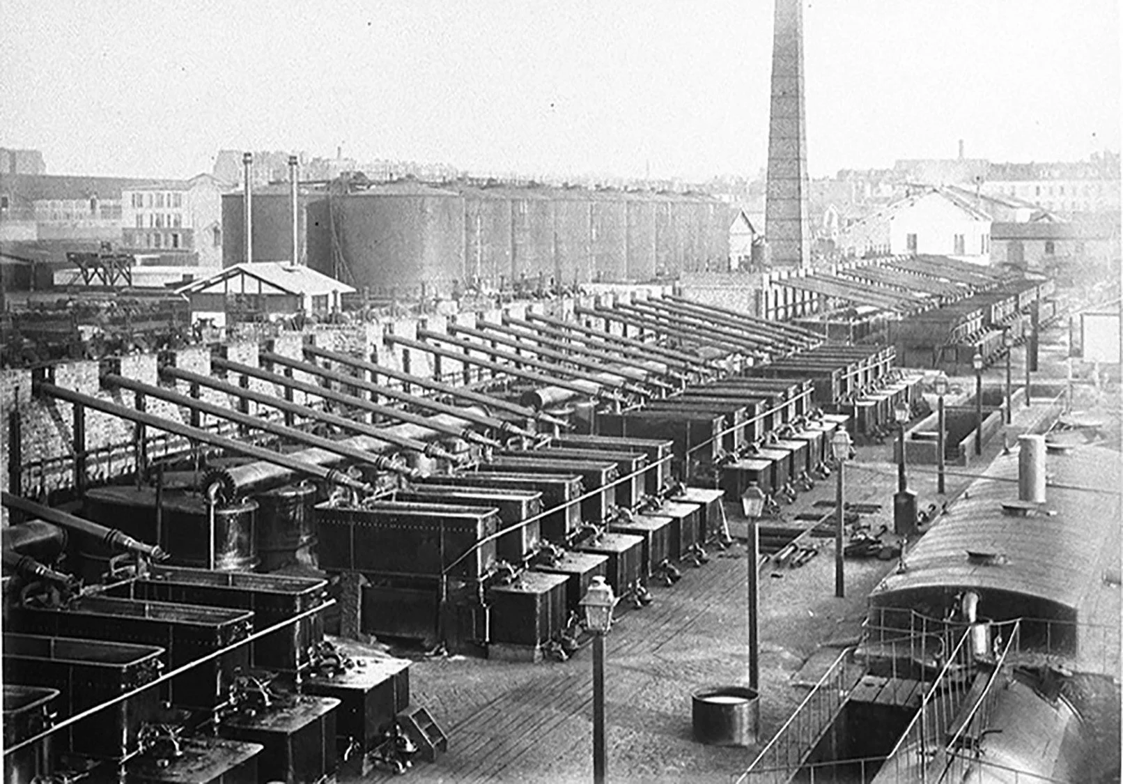 Black and white photo of an industrial waterfront with large tanks and pipes, possibly a shipyard or oil refinery, with buildings and smokestacks in the background.