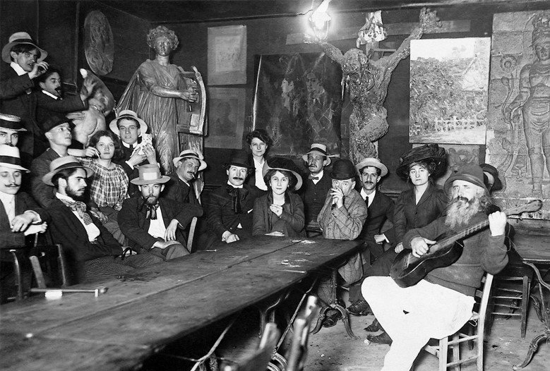 A vintage photograph of a group of people sitting around a large table in an art-filled room, with a man playing guitar at the right, and many of the people wearing period clothing and hats.