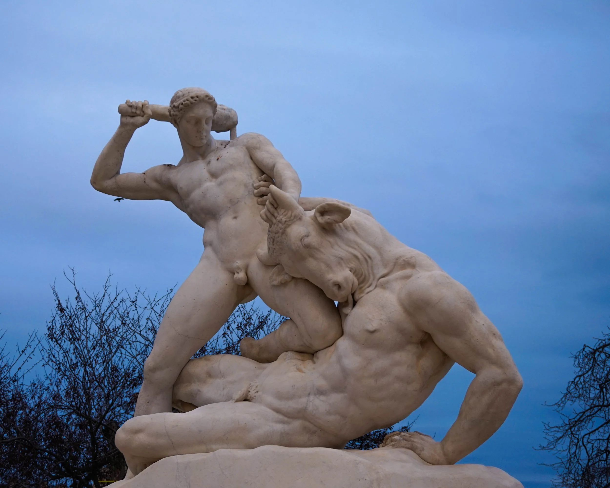 Marble sculpture of two muscular male figures engaged in a dynamic fight, set against a cloudy sky with leafless trees in the background.