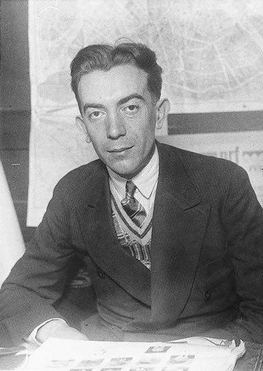 A young man in a suit and tie sitting at a desk with papers, in front of a chalkboard or large paper with writing.