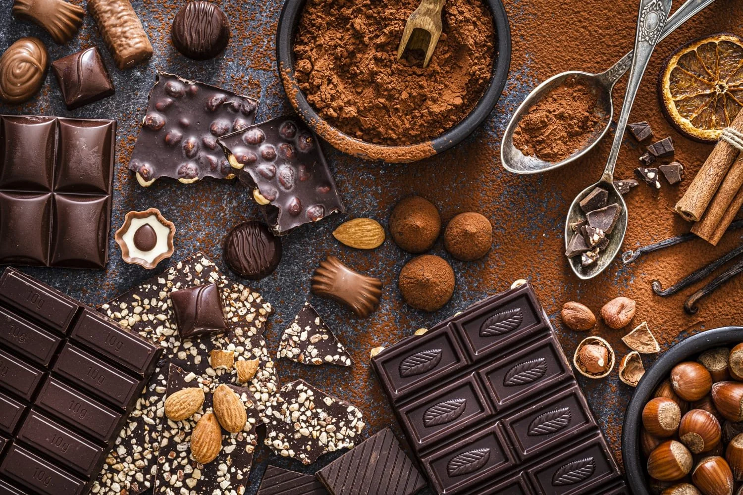 A variety of chocolates, cocoa powder, cinnamon sticks, dried lemon slice, nuts, and spices spread out on a dark surface for making or serving chocolates.