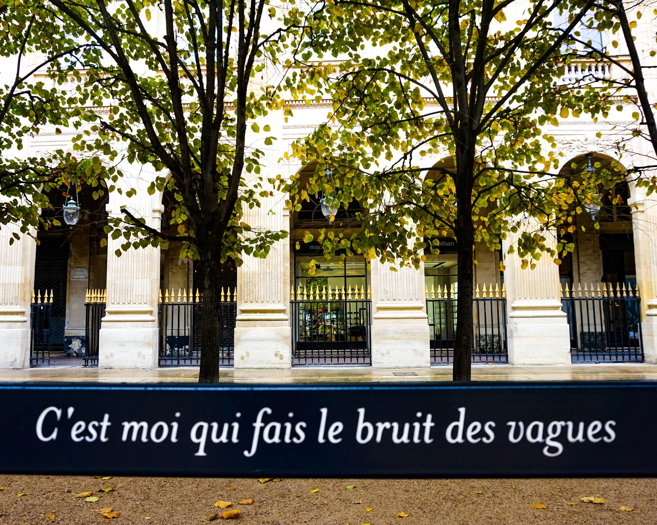 A building with tall columns and trees in front, with a French quote on a dark blue sign at the bottom reading 'C'est moi qui fais le bruit des vagues'.