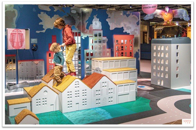 Two children playing on a colorful indoor city-themed play area with miniature buildings and a backdrop of blue trees and sky, in a children's museum or play center.