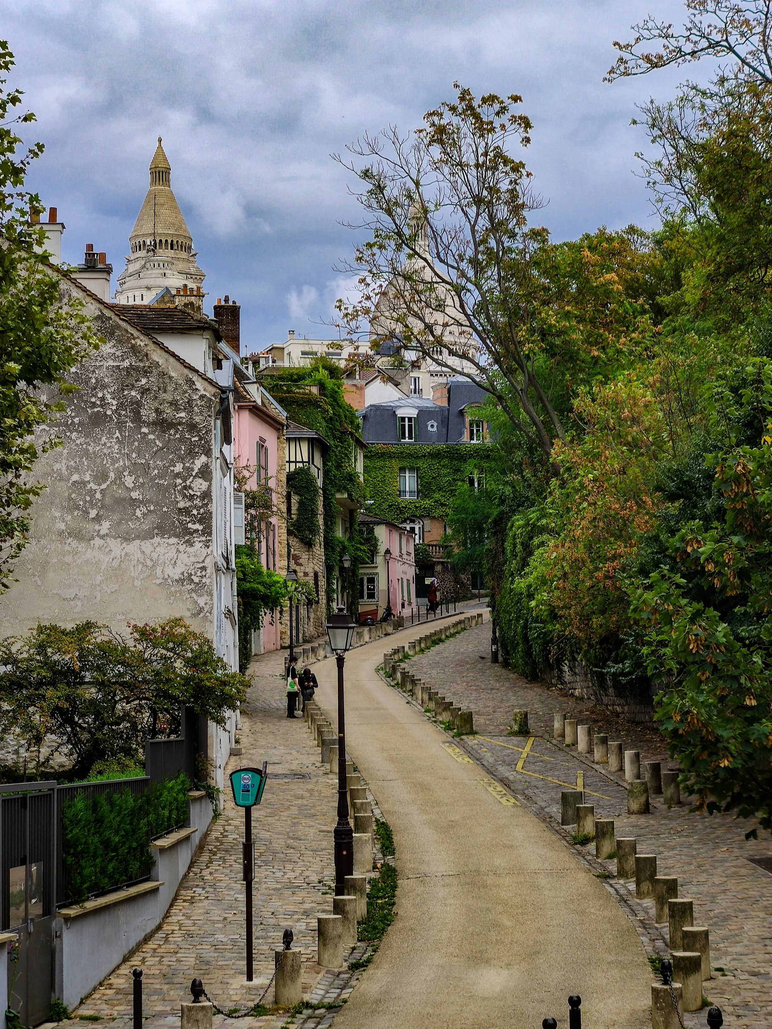 A street in Paris with a cobblestone sidewalk, trees on the right, pastel-colored houses on the left, and the Sacré-Cœur basilica visible in the background against a cloudy sky.
