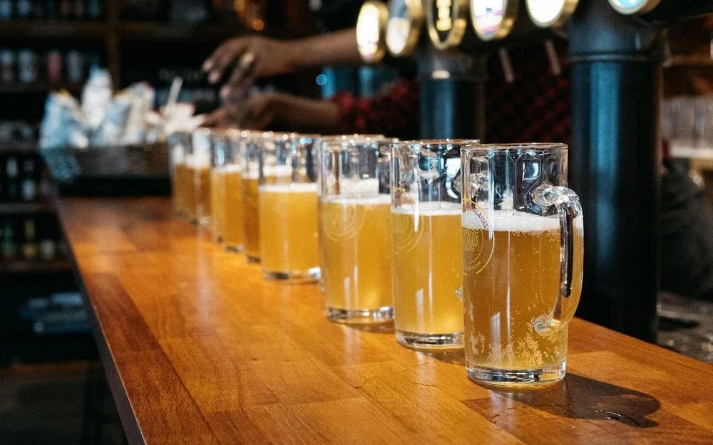 Six glasses of cloudy beer on a wooden bar counter in a brewery or bar setting.