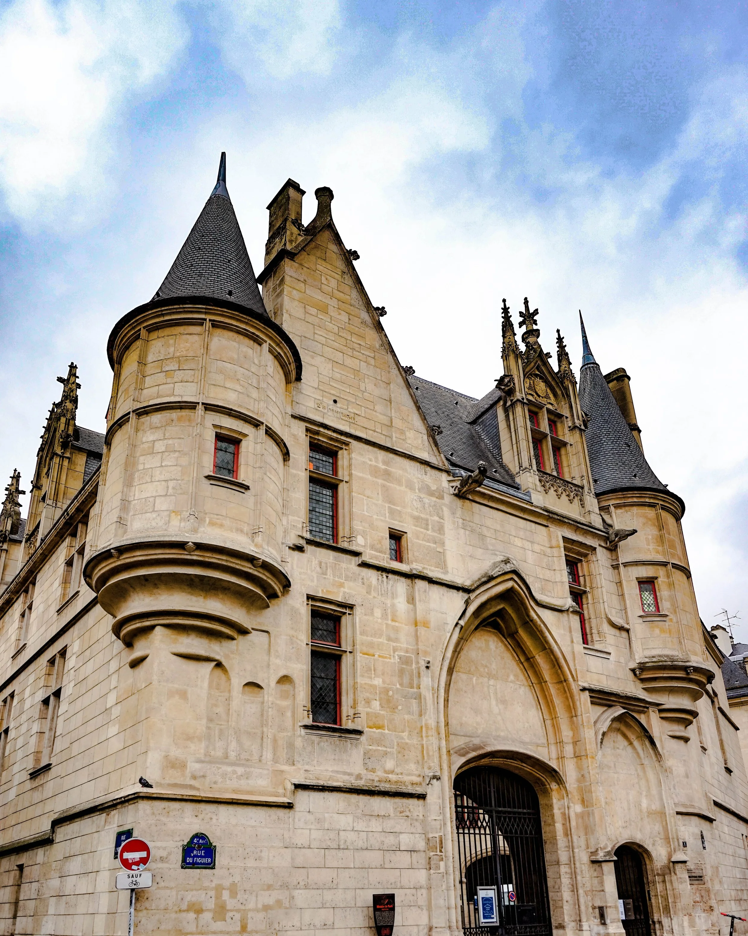 A historic castle or chateau with Gothic architecture, featuring turrets with conical roofs, intricate stonework, and narrow windows, under a partly cloudy sky.