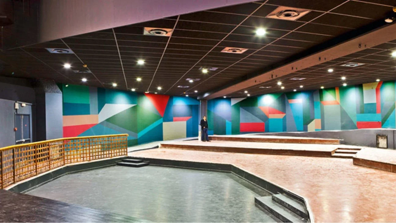 Empty indoor stage with colorful geometric mural backdrop and tiered steps, in a dimly lit room.