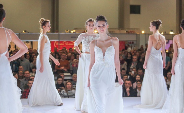 Models walking down the runway at a fashion show, wearing white dresses with an audience watching in the background.