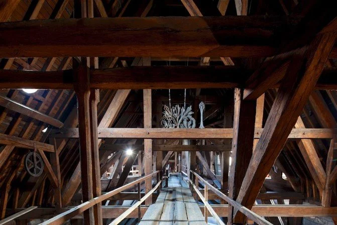 View of the interior of a wooden attic with exposed beams, a chandelier hanging from the ceiling, and a narrow walkway with railings.