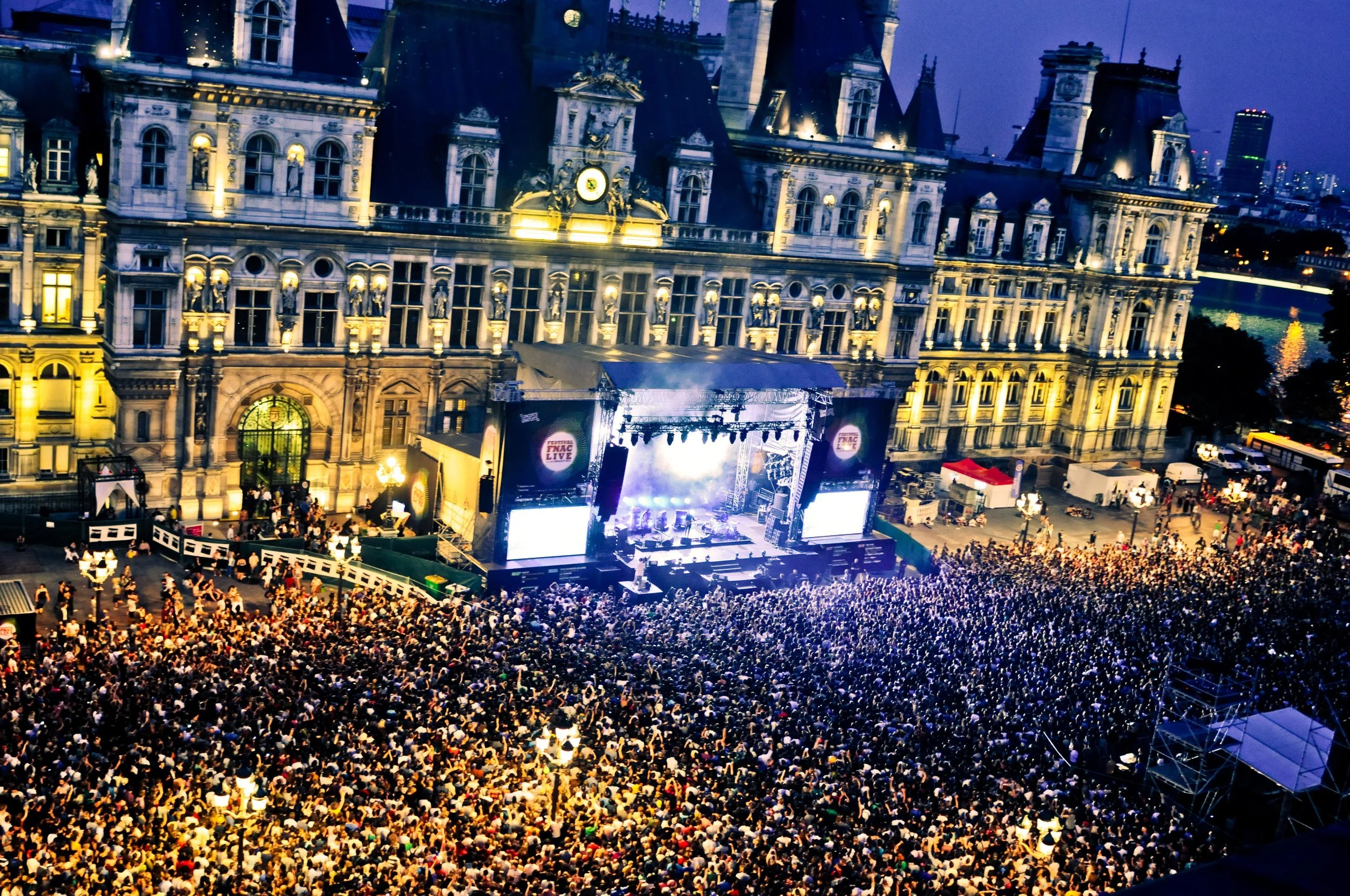 Large outdoor concert at night with a crowd in front of a stage set against a historic, illuminated building.