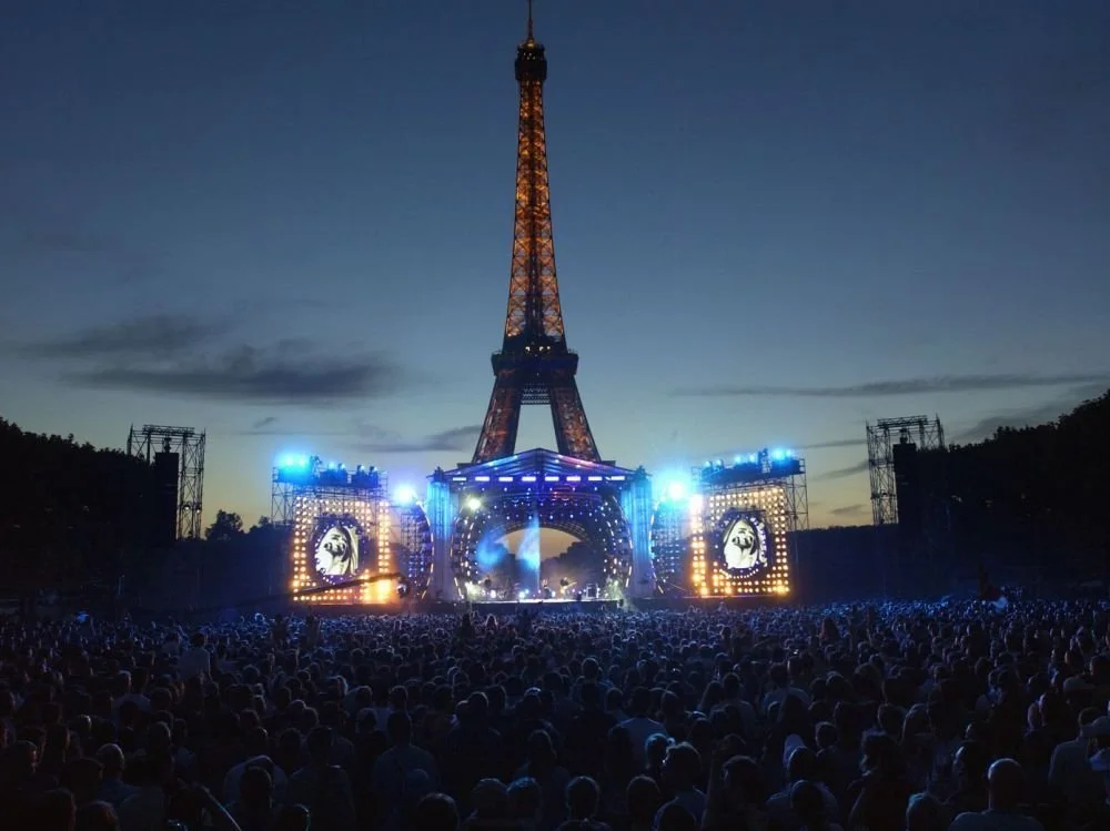Nighttime concert in front of the Eiffel Tower with a large crowd and stage lighting.