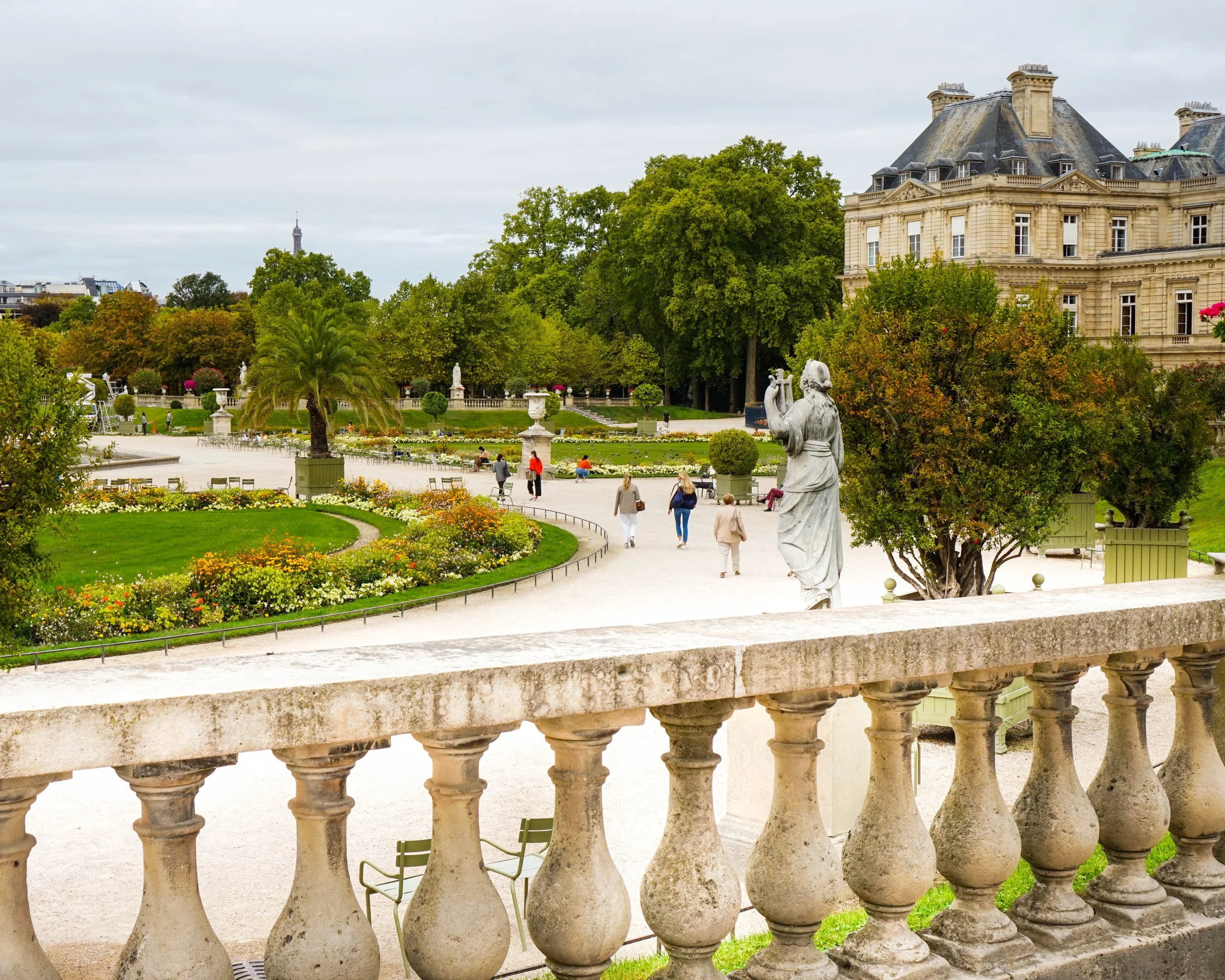 A park with a stone statue of a woman playing a musical instrument, lush green trees, colorful flower beds, people walking, and a large historic building with a gray roof in the background.