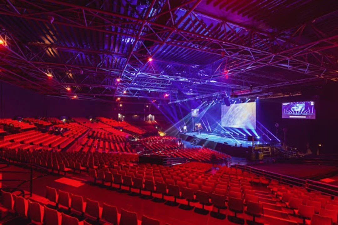 Empty auditorium with red seats, stage with blue and purple lighting, large screen, and overhead truss system with lights.